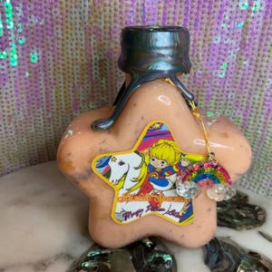 Peut inclure: Une bouteille en forme d'étoile contenant un liquide rose et un charme arc-en-ciel. La bouteille porte une étiquette indiquant "Starlite Sparkle Magic Potion Bottle".