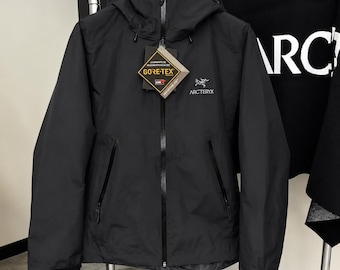 Op de afbeelding: Een zwarte Arc'teryx jas met capuchon, voorzien van een volledige ritssluiting aan de voorzijde, twee zijzakken met rits en verstelbare manchetten. Een Gore-Tex label hangt aan de voorzijde en het logo is wit op de borst.