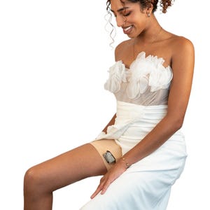 Op de afbeelding: Een vrouw die een witte strapless jurk draagt met een gerimpelde buste en een witte rok. Ze zit met haar benen gekruist en heeft een klein apparaat aan haar bovenbeen bevestigd.