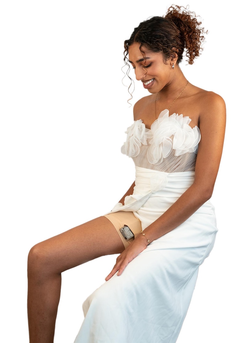 Op de afbeelding: Een vrouw die een witte strapless jurk draagt met een gerimpelde buste en een witte rok. Ze zit met haar benen gekruist en heeft een klein apparaat aan haar bovenbeen bevestigd.