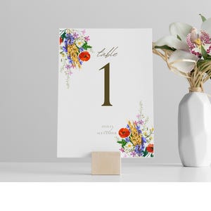 May include: A white table number card with the number 1 in dark green, the word "table" in script, and floral accents. The card is propped up by a small wooden block. A white vase with flowers is in the background.