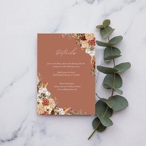 May include: A terracotta-coloured invitation card featuring floral accents and the word "Details" in elegant script. The card is adorned with a botanical design of white, orange, and brown flowers. A sprig of green eucalyptus adds a natural touch.