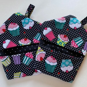May include: Two black and white pot holders with a polka dot pattern and a cupcake design. The pot holders have a loop handle.