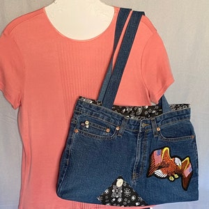 May include: A blue denim tote bag with a black and white bandana lining and an embroidered eagle patch. The bag has two denim straps.