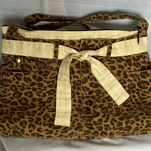 May include: A brown and tan leopard print tote bag with a cream colored fabric belt and bow. The bag has a top zipper closure and a brown fabric lining.