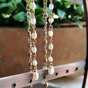 May include: Gold-toned earrings with dangling white pearls. The earrings are displayed on a copper-colored jewelry stand against a blurred green background. The earrings have a delicate chain design.