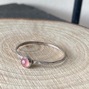 May include: A delicate silver ring with a pink gemstone set in the center. The ring is hammered for a textured look.