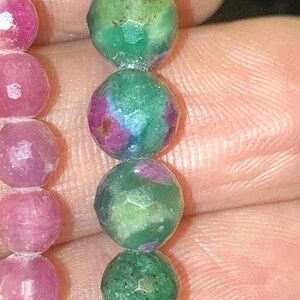 May include: Close-up of two beaded bracelets. One bracelet features faceted pink beads, while the other showcases green beads with red and blue inclusions. The beads are round and appear to be gemstones.