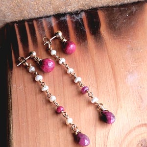 May include: A pair of gold-toned earrings with a stud and a long, dangling design. The earrings feature small white pearls, round and teardrop-shaped pink gemstones. The earrings are displayed on a wooden surface.