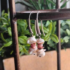 May include: A pair of dangle earrings with silver hooks. Each earring features a cluster of white, irregular-shaped pearls and pink gemstones. The earrings are displayed against a backdrop of green foliage.