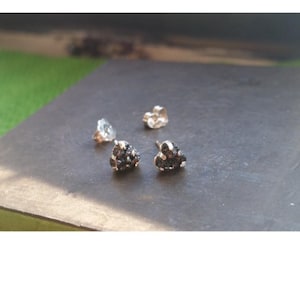 May include: Close-up of four stud earrings. Two earrings have a cluster of small, dark, textured stones. The other two earrings have a clear, faceted stone and a gold-colored setting. The earrings are on a dark, textured surface.