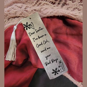 Pode incluir: Marcador de livros de metal prateado com borla, com a frase "Dear Santa, I've been a Good Girl, send me your Bad Boys." Inclui ilustrações de flocos de neve e caixas de presente.