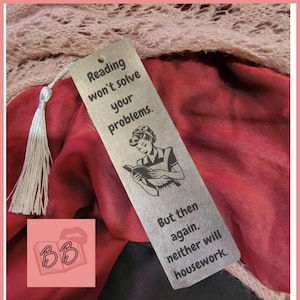 Puede incluir: Un marcapáginas plateado con una ilustración en blanco y negro de una mujer leyendo un libro. El marcapáginas tiene el texto "Reading won't solve your problems. But then again, neither will housework."