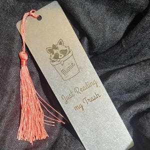 May include: A silver bookmark with a raccoon illustration in a bucket, the name "Marie", and the text "Just Reading my Trash". It has a coral tassel attached to the top. The bookmark is rectangular.