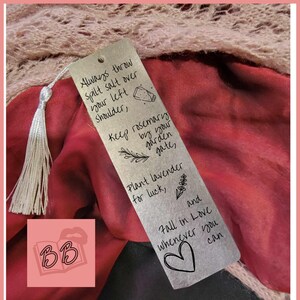 May include: A silver bookmark with black text and illustrations. The bookmark features a quote about throwing salt over your left shoulder, planting rosemary by your garden gate, and planting lavender for luck. The bookmark also includes a heart and the text "Fall in Love whenever you can."