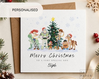 May include: A square greeting card featuring a watercolor illustration of Toy Story characters around a Christmas tree. Text reads: Merry Christmas TO A VERY SPECIAL SON Elijah. The card is set against a brown envelope with pine branches.