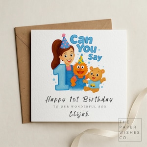 May include: A birthday card with a brown envelope. The card features a cartoon illustration of a girl, a number 1, and two cartoon characters. Text reads "Can You Say Happy 1st Birthday to our wonderful son Elijah."