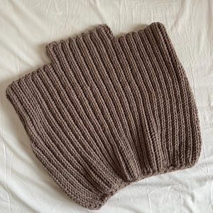 May include: Two brown knitted neck warmers are shown on a white surface. The neck warmers are made of a thick, ribbed knit fabric. The colour is a muted brown, and the texture of the knit is clearly visible.