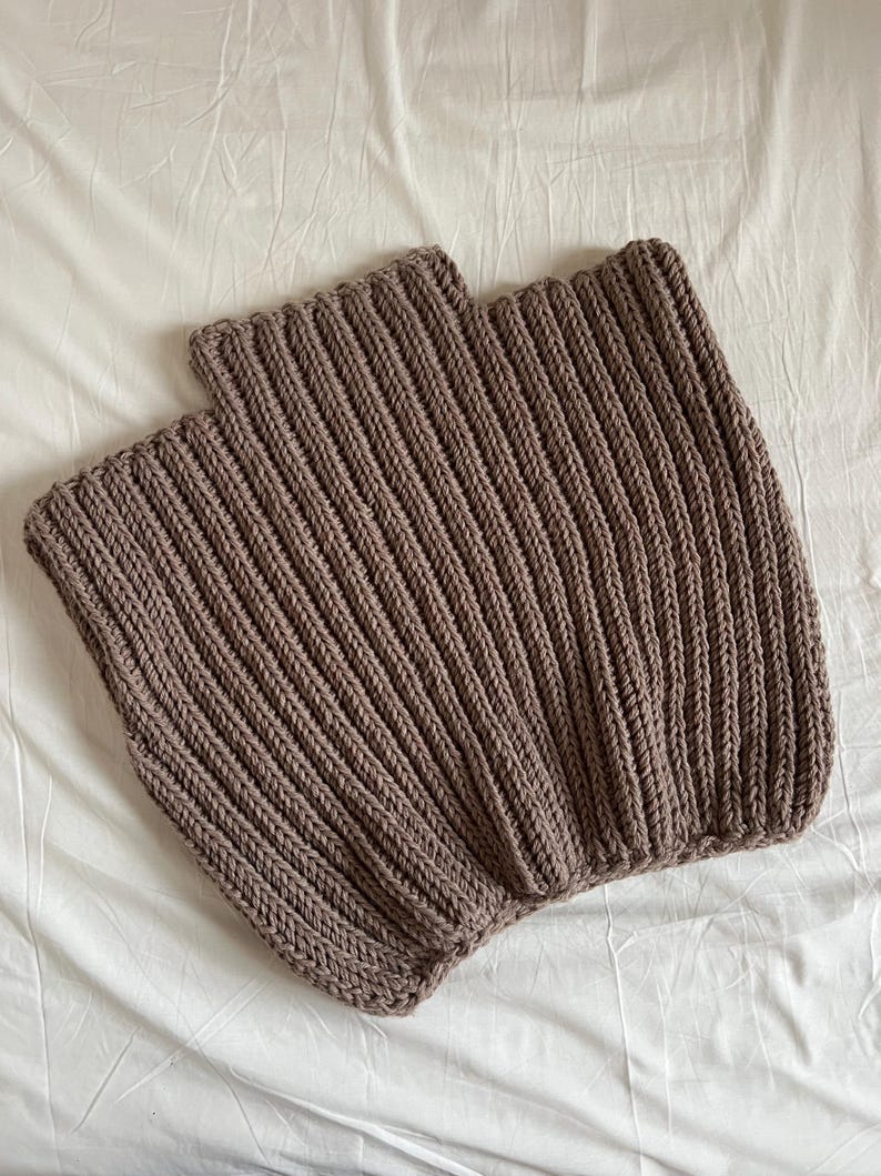 May include: Two brown knitted neck warmers are shown on a white surface. The neck warmers are made of a thick, ribbed knit fabric. The colour is a muted brown, and the texture of the knit is clearly visible.