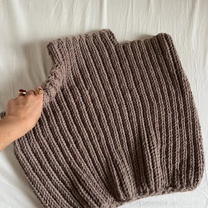 May include: A knitted top in a taupe hue, featuring a ribbed design. The garment has a high neckline and a square shape. The thick knit gives a textured look. The top is shown on a white surface.