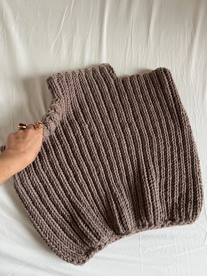 May include: A knitted top in a taupe hue, featuring a ribbed design. The garment has a high neckline and a square shape. The thick knit gives a textured look. The top is shown on a white surface.