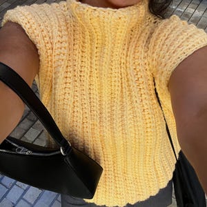 May include: A yellow crochet top with a high neckline and short sleeves. The top is made of a thick, textured yarn. A black handbag with a zip is visible on the left side of the image.