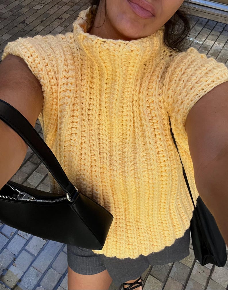 May include: A yellow crochet top with a high neckline and short sleeves. The top is made of a thick, textured yarn. A black handbag with a zip is visible on the left side of the image.