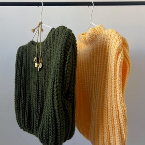 May include: Two chunky knit sweaters hang on a black rail. One is olive green, the other is a soft yellow. A gold-toned necklace with leaf-shaped pendants hangs from the green sweater. The sweaters are on white metal hangers.