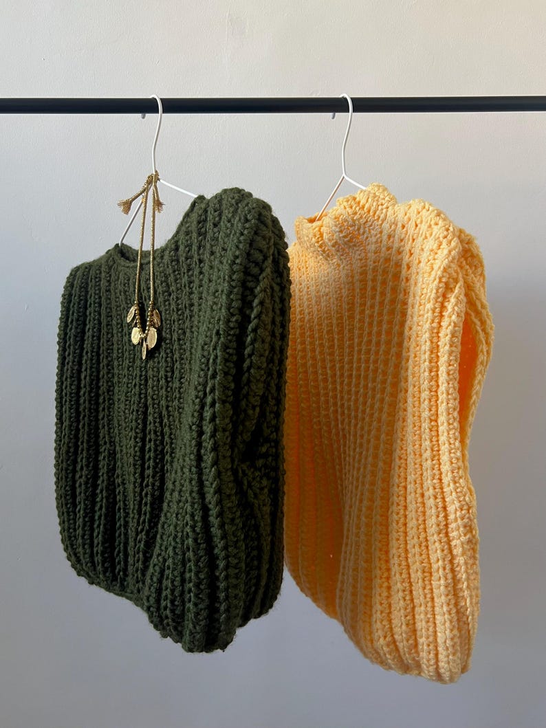 May include: Two chunky knit sweaters hang on a black rail. One is olive green, the other is a soft yellow. A gold-toned necklace with leaf-shaped pendants hangs from the green sweater. The sweaters are on white metal hangers.