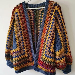 Peut inclure: Un cardigan en crochet multicolore, de style granny square, sans fermeture. Il présente un assemblage de carrés bleus, jaunes, oranges, rouges et bruns, avec une bordure et des poignets bleus unis. Le vêtement est suspendu à un cintre blanc.