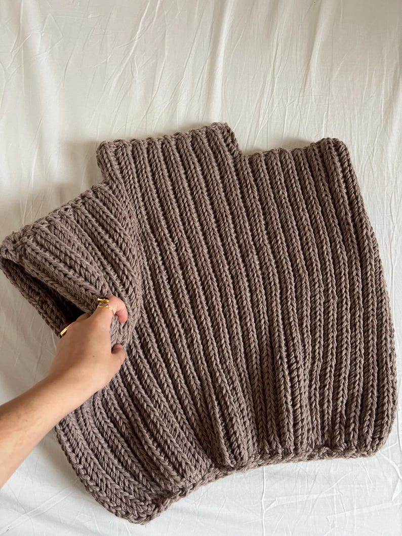May include: A close-up of a chunky knit, taupe-coloured sweater. The sweater is made with thick yarn and features a ribbed design. The sleeves are folded over. The sweater is laying on a white surface.