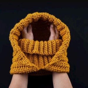 Op de afbeelding: Een mosterdgele gehaakte cowl met een capuchon. De cowl is dubbelgevouwen en wordt vastgehouden door twee handen.
