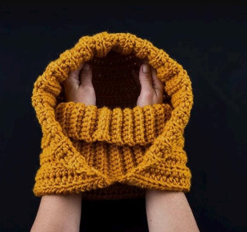 Op de afbeelding: Een mosterdgele gehaakte cowl met een capuchon. De cowl is dubbelgevouwen en wordt vastgehouden door twee handen.