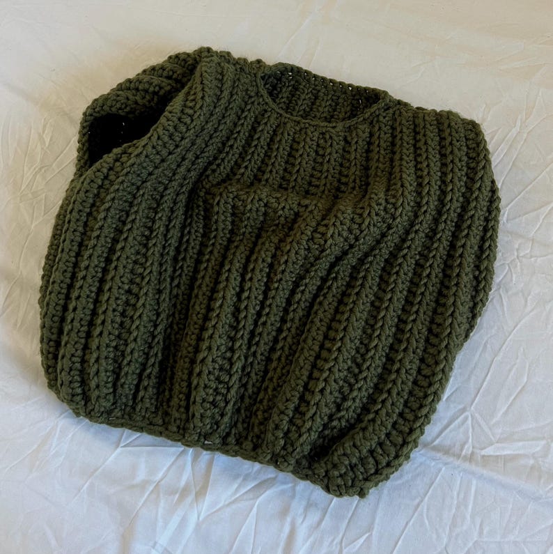 May include: An olive green, hand-knitted sweater. The sweater has a ribbed design across the body and sleeves. The neckline is a simple crew neck. The garment is made of a thick, textured yarn, giving it a cosy appearance.