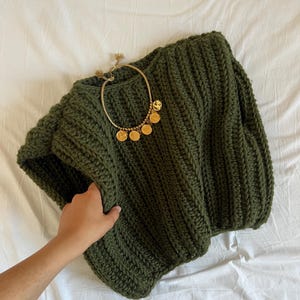 Può includere: Un maglione lavorato a maglia grossa verde oliva con una collana dorata con pendenti a moneta. Il maglione ha una trama a coste e uno scollo ampio. La collana è drappeggiata sul maglione.