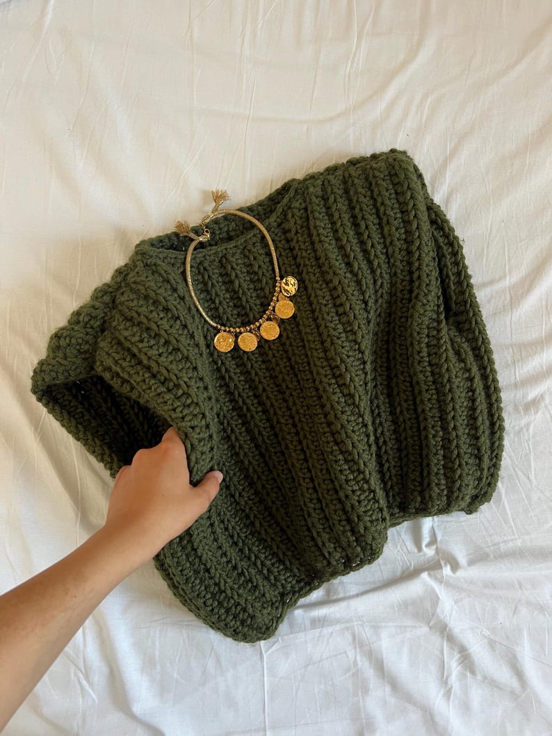 May include: An olive green, chunky knit jumper with a gold-toned necklace featuring coin pendants. The jumper has a ribbed texture and a wide neckline. The necklace is draped over the jumper.