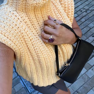 May include: A pale yellow crochet top with a high collar and a black purse. The image shows the texture of the crochet and the design of the top. Several rings are visible on the hand. The top is sleeveless.