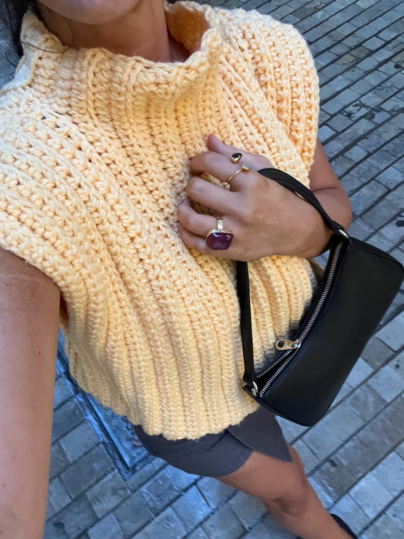 May include: A pale yellow crochet top with a high collar and a black purse. The image shows the texture of the crochet and the design of the top. Several rings are visible on the hand. The top is sleeveless.