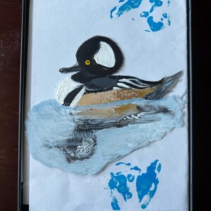 May include: A framed painting of a Hooded Merganser duck. The duck has a black head with a white patch, a white chest, and a brown and grey body. It is depicted on a blue water background with blue handprints.