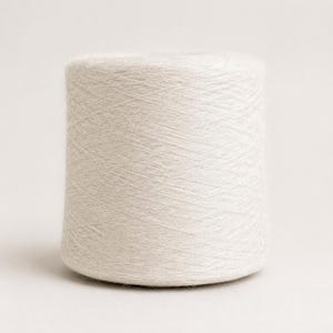 May include: A cylindrical spool of off-white yarn featuring a fine, soft texture. The thread is wound tightly in a cross-hatch pattern around the central core, creating a uniform, smooth surface across the entire exterior of the yarn cone.