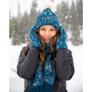 May include: A matching winter set featuring a blue knit hat with a faux fur pom, mittens, and a scarf with fringe. The set is a mix of blue and white yarn, perfect for cold weather. The person in the image is wearing a black jacket.