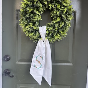 May include: A green wreath with a full, leafy appearance hangs on a dark green door. A white sash with an embroidered floral "S" is tied to the wreath. The door has a black handle and a small window at the top.