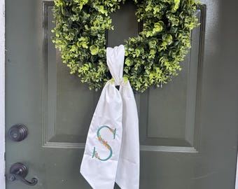 Puede incluir: Una corona verde con una apariencia frondosa completa cuelga de una puerta verde oscuro. Una faja blanca con una "S" floral bordada está atada a la corona. La puerta tiene una manija negra y una pequeña ventana en la parte superior.