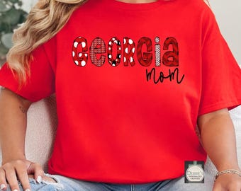 Puede incluir: Una camiseta roja de manga corta con la palabra GEORGIA en letras grandes estampadas con varios diseños en rojo y negro, incluyendo lunares y cuadros. Debajo de la palabra, el término mom está impreso en una fuente cursiva negra sencilla.
