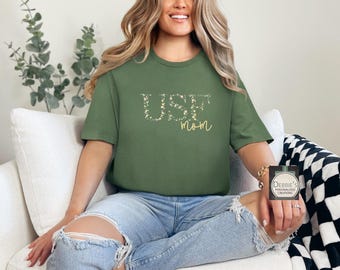 Puede incluir: Una camiseta verde con las palabras "Use Mom" en letras florales. La camiseta la lleva puesta una persona con el pelo largo y rubio.