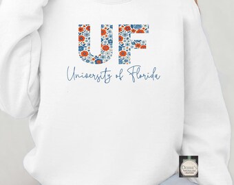 Puede incluir: Sudadera blanca con las letras "UF" en un diseño floral de flores azules y naranjas. Debajo, las palabras "University of Florida" están escritas en una fuente cursiva. Sudadera de estilo casual.