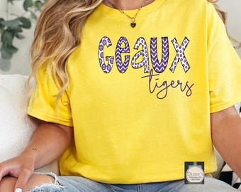 Puede incluir: Una camiseta amarilla brillante de manga corta con la palabra GEAUX en letras grandes de color morado y blanco con un contorno negro cosido, seguida de la palabra tigers en cursiva morada. Un pequeño logotipo cuadrado es visible en la parte inferior.
