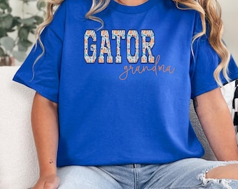 Puede incluir: Una camiseta de manga corta de color azul real con la palabra GATOR en letras de bloque con un patrón floral multicolor, y la palabra grandma escrita en letra cursiva naranja debajo del texto en bloque.