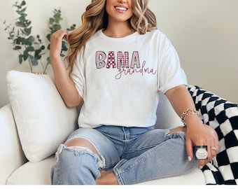 Puede incluir: Una camiseta blanca de manga corta con el texto BAMA en un patrón de cuadros y lunares rojos y blancos, con la palabra grandma escrita en cursiva roja debajo. La camiseta se combina con unos vaqueros azul claro desgastados.