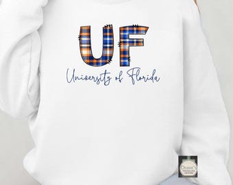Puede incluir: Sudadera blanca con las letras "UF" en un diseño a cuadros azul y naranja. Debajo, la frase "University of Florida" en azul marino. Cuello redondo.